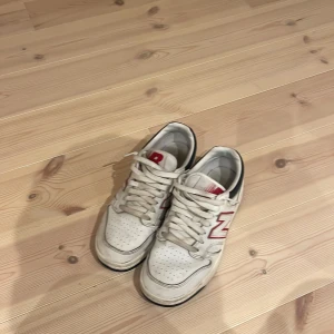 Vita sneakers från New Balance - Säljer ett par vita New Balance sneakers med röda detaljer och svart häl. Skorna har snörning och en klassisk rund tå. Perfekta för en avslappnad stil.