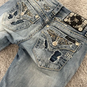Blå jeans från Miss Me - Snygga blå jeans från Miss Me med unika broderade detaljer och slitningar. Byxorna har en bootcut-stil och är dekorerade med knappar och mönster på bakfickorna. Perfekta för dig som vill ha något extra i garderoben! i jättebra skick!