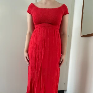 Röd klänning med smockdetaljer - Snygg röd klänning med smockdetaljer upptill och korta ärmar. Klänningen har en lång och flödande design som ger en elegant look. Perfekt för sommarens alla tillfällen. Endast använd fåtal gånger.