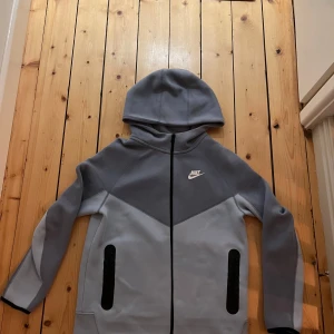  nike tech blå tröja  - Säljer en snygg grå och blå huvtröja från Nike med dragkedja och fickor. Perfekt för en sportig look. Tröjan har en justerbar huva och långa ärmar.