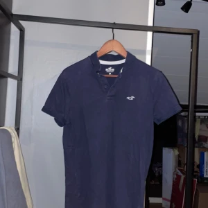 Mörkblå pikétröja från Hollister - Säljer en stilren mörkblå pikétröja från Hollister med korta ärmar och klassisk krage. Tröjan har en broderad logotyp på bröstet och är tillverkad i ett stretchigt material för extra komfort. Perfekt för en avslappnad look.