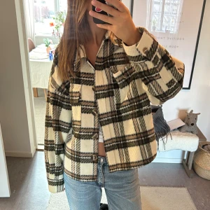 Rutig overshirt i beige och svart - Säljer en snygg rutig overshirt i beige och svart. Jackan har en klassisk krage och knäppning framtill. Perfekt för lager-på-lager-stil och ger en avslappnad look.