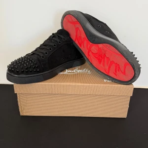 Svarta sneakers med nitar från Christian Louboutin - Snygga svarta sneakers i mocka från Christian Louboutin med coola nitar på tån. Skorna har en klassisk snörning och den ikoniska röda sulan. Perfekta för att ge din outfit en edgy touch.