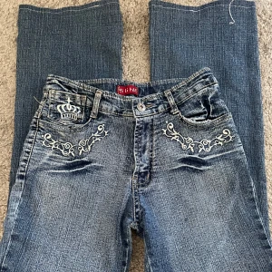 Low waist bootcut jeans - Snygga blå jeans med unika broderade detaljer av en krona och mönster både fram och bak. Säljer då dom är för korta💕 innerbenslängd:66cm, midjemått ca 64cm. Har justerbar sak vid midjan, skulle säga att den passar som xs