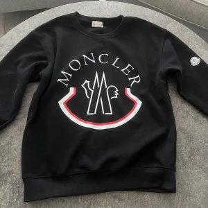 Svart sweatshirt från Moncler - Snygg svart sweatshirt från Moncler med stort tryck på framsidan. Tröjan har rund hals och ribbade muddar vid ärmslut och nederkant. Pris kan diskuteras!