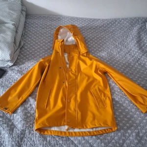 Gul regnjacka från Cross Sportswear - Säljer en snygg gul regnjacka från Cross Sportswear. Jackan har en huva och stängs med knappar och dragkedja. Perfekt för regniga dagar och håller dig torr och bekväm. Den har en vit insida och praktiska fickor framtill. Har några få fläckar, knappast märkbara 
