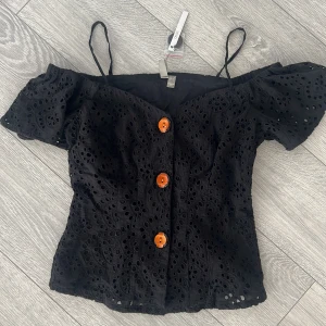 Svart offshoulder blus från ASOS - Säljer en snygg svart offshoulder blus från ASOS med orange knappar framtill. Blusen har ett vackert broderat mönster och smala axelband. Perfekt för en somrig look!