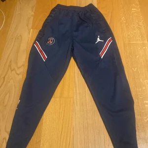 Mörkblå PSG träningsbyxor - Snygga mörkblå träningsbyxor från Jordan med PSG-logga. Byxorna har elastisk midja och vita detaljer med röd och vit rand. Perfekta för fotbollsträning eller avslappnad stil.