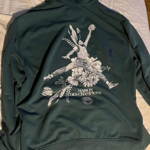 Grön hoodie från Jordan  - Snygg grön hoodie från Jordan med en unik design på ryggen som föreställer en figur i rörelse omgiven av blommor. Framsidan har en diskret logga på bröstet. Perfekt för en avslappnad stil.