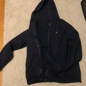 Mörkblå hoodie från Lyle & Scott - Säljer en stilren mörkblå hoodie från Lyle & Scott Junior. Den har en dragkedja framtill och en liten gul logga på bröstet. Perfekt för en avslappnad look.