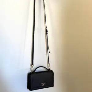 Elegant svart axelväska från Michael Kors med justerbar axelrem och guldfärgade detaljer. Väskan har en praktisk storlek och är perfekt för att bära dina viktigaste tillhörigheter. Den har en stilren design med ett litet handtag och logotyp framtill. Insidan innehåller flera platser för kort och ett fack med dragkedja. På utsidan finns ett fack för att förvara din mobiltelefon på ett säkert sätt. Ta bort axelremmen så får du en snygg clutch bag. Mått: B: 18,5 cm H: 11 cm D: 6,5 cm