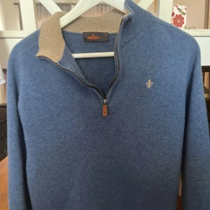 Blå tröja med dragkedja från Morris - Säljer en stilren blå tröja från Morris med dragkedja och en liten broderad detalj på bröstet. Tröjan har en kontrasterande beige krage och långa ärmar. Perfekt för en avslappnad men snygg look. Krympt lite så passar S bättre och har små skador mellan armarna