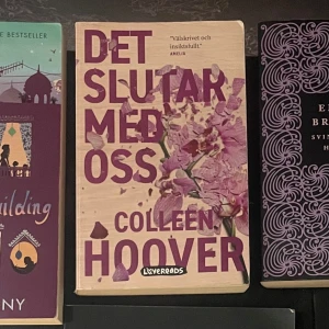 Det slutar med oss - Boken 'Det slutar med oss' av Colleen Hoover, utgiven av Lovereads. Omslaget är beige med lila text och illustrationer av blommor. En känslosam och insiktsfull berättelse som har blivit en bästsäljare.