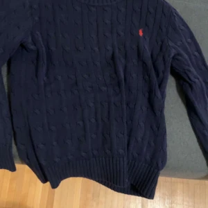 Grå kabelstickad tröja från Polo Ralph Lauren - Säljer en stilren grå kabelstickad tröja från Polo Ralph Lauren med det klassiska logotypbroderiet på bröstet. Tröjan har långa ärmar och ribbade muddar vid ärmslut och nederkant. Perfekt för en elegant och avslappnad look HELT NY ANVÄND 2 gånger tvättad 1-2 gånger 899 styck
