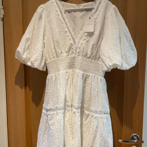 Vit klänning med puffärmar från By Malina - Charmig vit klänning från By Malina med puffiga kortärmar och v-ringad hals. Klänningen har ett fint prickigt mönster och spetsdetaljer. Den elastiska midjan ger en smickrande passform. Perfekt för sommarens alla tillfällen!