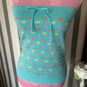 Turkos tubtopp med prickigt mönster - Säljer en söt turkos tubtopp med färgglada prickar i rosa, gult och orange. Toppen har en knytning framtill för extra detalj. Perfekt för en färgglad look! Armhåla till armhåla 33 cm , längd 37 Färgen är kanske mer turkos än blå
