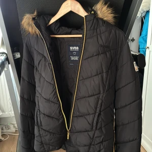 Svart quiltad jacka från Svea - Snygg svart quiltad jacka från Svea med dragkedja i guld och huva med fuskpäls
