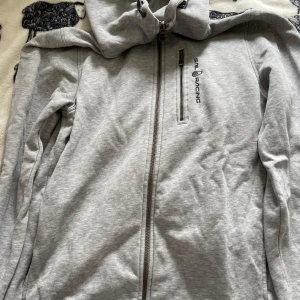Grå huvtröja från Sail Racing - Säljer en grå huvtröja från Sail Racing med dragkedja och en bröstficka. Tröjan har långa ärmar och en bekväm huva. Perfekt för en avslappnad stil. Dock är den lite sliten på ena ärmen vilket man kan se på bild men det går att sy 