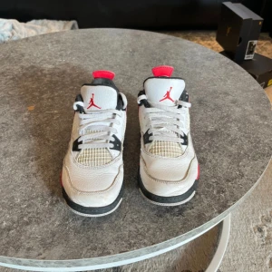 Jordens 4 - Säljer ett par jordens 4 med vit ovandel i skinn, svarta och röda detaljer samt meshpaneler på sidorna. Skorna har snörning och rund tå, perfekt för dig som gillar streetstyle.