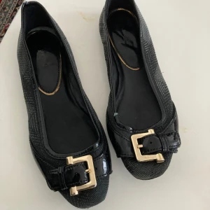 Svarta ballerinaskor från Burberry med spänne.storlek 38 - Säljer ett par svarta ballerinaskor från Burberry med elegant guldfärgat spänne framtill. Skorna har en diskret textur i materialet och lackade detaljer vid tån. Perfekta för dig som vill ha en klassisk och stilren look.