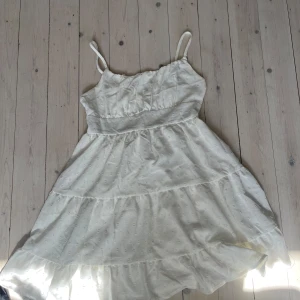 Vit sommarklänning med smala axelband - Söt vit klänning med smala, justerbara axelband och lätt volang i kjolen. Klänningen har resår i ryggen och är sydd i ett tunt, luftigt tyg med diskret struktur. Perfekt för varma dagar och har en romantisk känsla. Ens axelbandet är av med funkar med en säkerhetsnål 