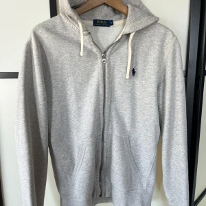 Grå hoodie med dragkedja från Polo Ralph Lauren - Säljer en ljusgrå hoodie från Polo Ralph Lauren med dragkedja och två fickor framtill. Tröjan har huva med vita snören och en liten broderad logga i marinblått på bröstet. Självklart äkta!