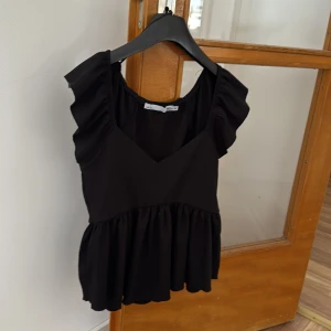 Topp  - Säljer en svart topp med volangärmar och v-ringning. Toppen har en lös passform och är tillverkad i ett lätt, syntetiskt material. Perfekt för dig som gillar en feminin och trendig stil.