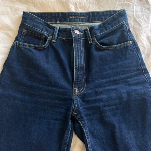Mörkblå jeans från Nudie Jeans - Säljer ett par klassiska mörkblå jeans från Nudie Jeans med raka ben och normal passform. Jeansen har fem fickor, gylf med dragkedja och Nudie Jeans signatursöm på bakfickan. Perfekta för en stilren vardagslook.