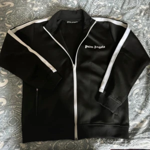 Palm Angels Zip-Up Kofta – Svart |  -  Säljer en snygg och stilren zip-up kofta från Palm Angels i klassisk svart färg. Ikonisk design med logga framtill och dragkedja hela vägen. Perfekt för dig som vill kombinera streetwear med lyxig känsla.