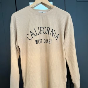 Beige ribbad långärmad tröja med tryck - Beige ribbad långärmad tröja med svart text 'California West Coast' på bröstet. Tröjan har rund halsringning och är gjord i ett mjukt, stretchigt material. Perfekt för en avslappnad och trendig look.