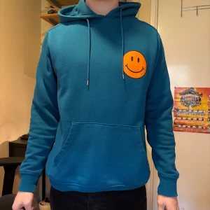 Blå hoodie med smiley  - Säljer en blå hoodie med huva och dragsnören. På bröstet finns ett orange smiley-tryck. Tröjan har en stor magficka och långa ärmar. Perfekt för en avslappnad stil.
