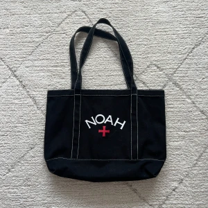 Svart tygväska från Noah - Säljer en svart axelväska från Noah med vit kontrastsöm och tryckt logga framtill. Väskan har dubbla handtag och är tillverkad i kraftigt tygmaterial. Perfekt för att bära med sig det viktigaste till vardags.