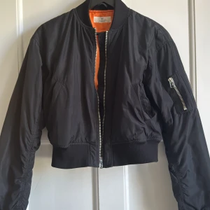 Svart bomberjacka från H&M - Säljer en svart bomberjacka från H&M med orange foder. Jackan har dragkedja framtill, fickor på sidorna och en ärmficka med dragkedja. Croppad modell med ribbade muddar vid ärmslut och midja.