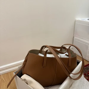 Brun handväska i skinn Poelene  - Säljer en stilren brun handväska i skinn med långa handtag och minimalistisk design. Perfekt för att bära dina vardagsprylar och passar till många olika outfits. Kommer i originalkartong. Original priset är 4200 kr