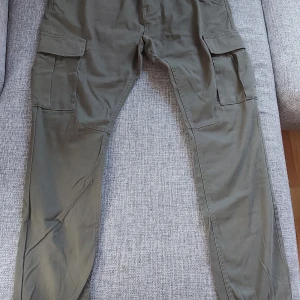 Ankel byxor  - Nya byxor, mått,: längd 98 cm mätt på framsidan, 101 cm mätt på baksidan, 39 cm höft till höft , innerbenslängd ca 73,5 cm. Fickor i fram och bak, en ficka på sidan på varje ben. Fickorna i bak och på sidorna har kardborreband som knäppning, hällor . Rökfritt men det finns katt.