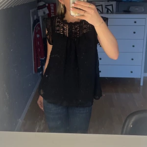 Svart blus med spetsdetaljer - Säljer en svart blus med vackra spetsdetaljer upptill och volang vid axlarna. Blusen är ärmlös och har en luftig passform, perfekt för varmare dagar.