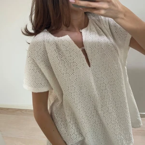 Vit blus med broderade detaljer - Säljer en luftig vit blus med korta ärmar och v-ringning med knyt framtill. Blusen har ett vackert broderat hålmönster över hela tyget och är perfekt för varma dagar.