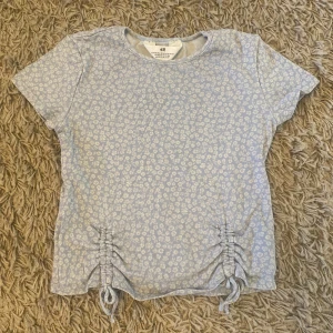 Blommig t-shirt  - Säljer en ljusblå t-shirt med vita små blommor och detaljer längst ner på framsidan. Perfekt nu för sommaren.