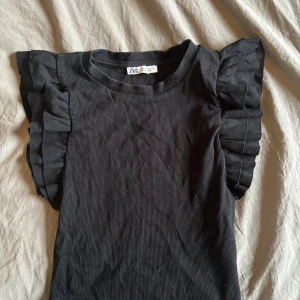 Svart ribbad topp med volangärmar från Zara  från Zara - Säljer en svart ribbad topp från Zara med snygga volangdetaljer på axlarna. 