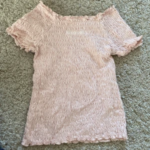 Ljusrosa smockad tröja från H&M - Säljer en ljusrosa smockad topp från H&M med korta ärmar och stretchigt material. Toppen har en söt, rynkad struktur och en feminin passform. Perfekt till sommaren och enkel att matcha med jeans eller kjol.