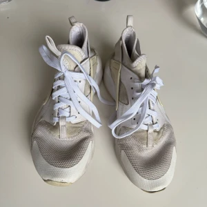 Vita Nike sneakers Air Huarache - Säljer ett par vita Nike Air Huarache sneakers med mesh och syntetdetaljer. Skorna har snörning, rund tå och en tjock, platt sula med AIR-logga bak. Perfekta för dig som gillar klassiska och sportiga sneakers. Men det finns ett litet märke som visas på bilden