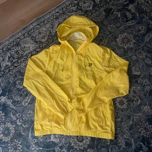 Gul vindjacka med huva från Lyle & Scott - Säljer en tunn gul vindjacka från Lyle & Scott med huva och dragkedja framtill. Jackan har två sidofickor och en liten logga på bröstet. Perfekt för blåsiga dagar och enkel att packa ner i väskan.