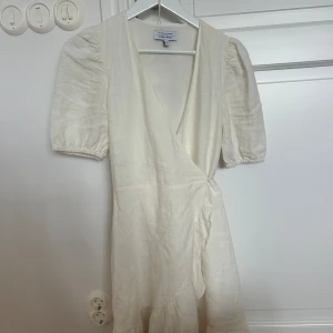 Vit omlottklänning från & Other Stories - Superfin vit omlottklänning från & Other Stories med puffärmar och knyt i midjan. Klänningen har v-ringning och volangdetalj nertill. Perfekt som student klänning eller konfirmationsklänning! Använd en gång