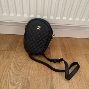 Svart quiltad axelväska  - Säljer en svart quiltad axelväska  med guldfärgade detaljer och klassisk logga framtill. Väskan har justerbar axelrem och elegant oval form. Perfekt för att bära det viktigaste med stil.