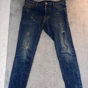 Blå slitna jeans från Dsquared2 - Säljer ett par blå jeans från Dsquared2 med slitningar och trasiga detaljer på benen. Klassisk femficksmodell med knappgylf och smal passform. Perfekta för en cool och avslappnad stil.