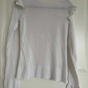 Vit volangtröja från H&M - Säljer en vit långärmad tröja från H&M med volangdetaljer på axlarna. Tröjan är ribbad och har en rund halsringning samt lätt utsvängda ärmslut. Perfekt för dig som gillar stilrena och feminina detaljer.