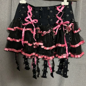 Kjol från Poizen S - Unik svart kjol med rosa snörning och volanger i flera lager. Kjolen har dekorativa metallringar och detaljer, samt svarta och rosa band som ger en edgy look. Perfekt statement piece för dig som vill sticka ut.