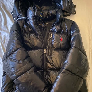 Svart pufferjacka från Polo Ralph Lauren - Säljer en svart glansig pufferjacka från Polo Ralph Lauren med huva och dragkedja. Jackan har en röd broderad logga på bröstet och hög krage. Perfekt för kyliga dagar och riktigt snygg streetstil. Säljs pga används inte längre😊 använd ca 4-6 ggr, finns inga märken eller nånting ser ut som helt ny jacka 🧥
