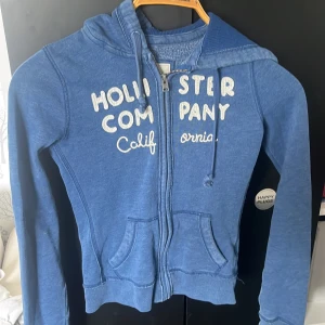 Blå hoodie från Hollister - Säljer en blå hoodie från Hollister med vit broderad text på bröstet och dragkedja framtill. Tröjan har huva med dragsko och två fickor framtill. Perfekt för en avslappnad stil.