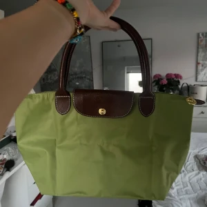 Grön axelväska  - Säljer en klassisk grön axelväska med bruna handtag och lock i skinn. Väskan har guldknapp och är perfekt för att bära dina vardagsprylar. Enkel och stilren design med plats för det viktigaste. Ny skick 